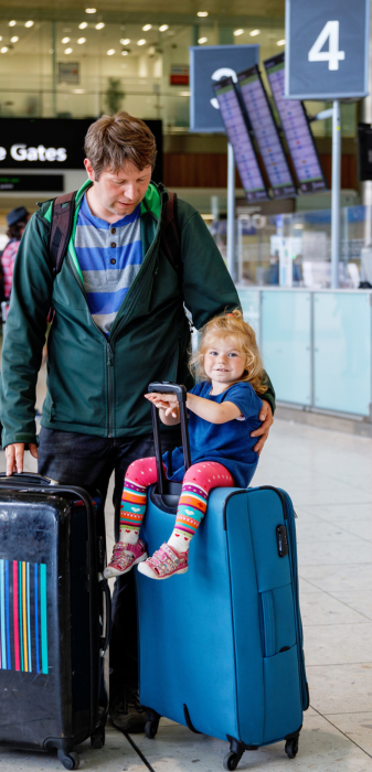 Un homme transporte 2 grandes valises dans un aéroport et un enfant est assis sur la valise de droite.